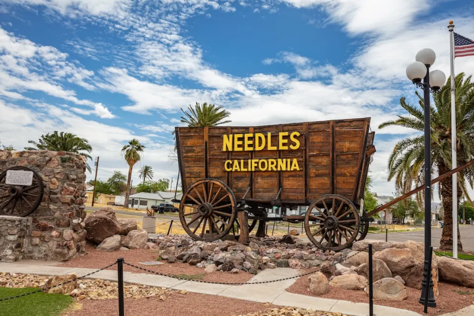 Sign for Needles, California installed on wooden wagon with residential neighborhood visible in background.